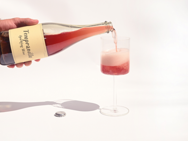 Hand pouring Swick Wines 2021 Tempranillo Sparkling Wine fizzy pink red wine from bottle into a glass with bubbles and bottle cap on beige background