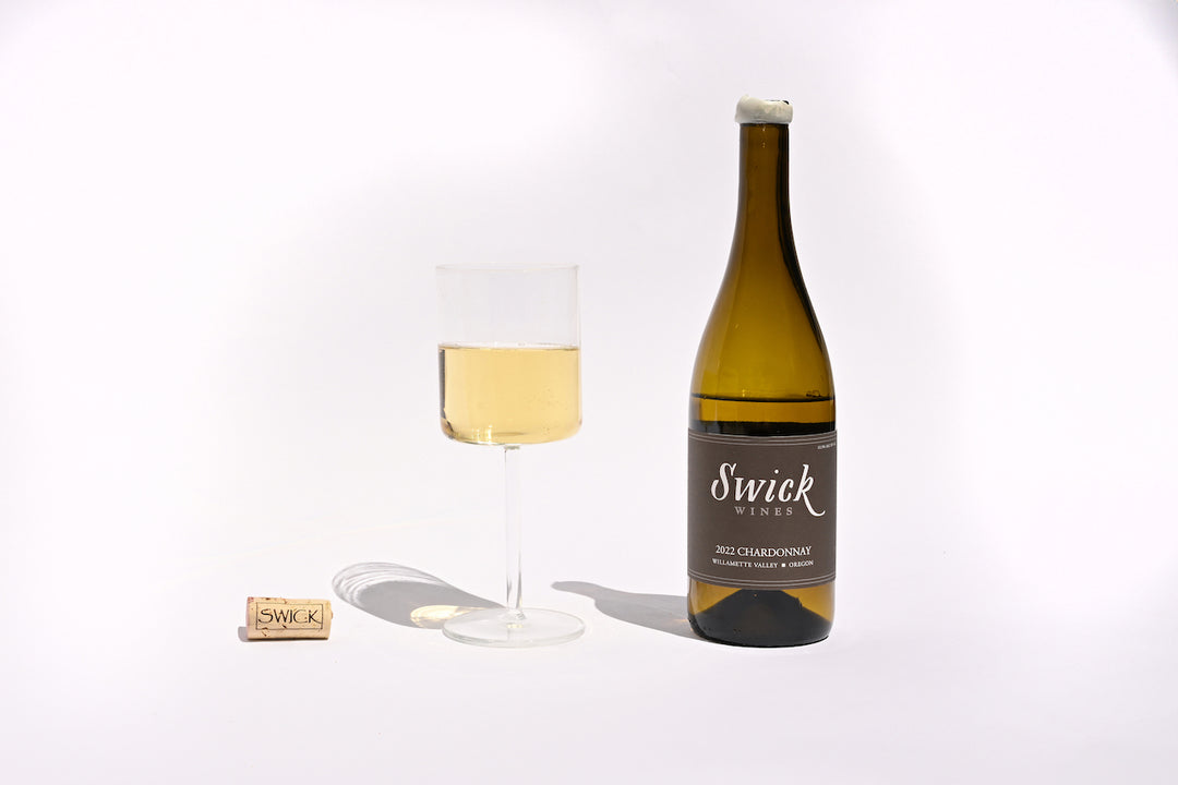 Swick Wines 2022 Chardonnay Willamette Valley dark green bottle with cream and black label, filled glass of pale yellow white wine, and cork on beige background