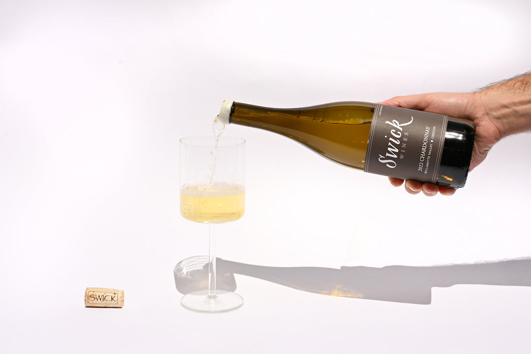 Hand pouring Swick Wines 2022 Chardonnay Willamette Valley pale yellow white wine from dark green bottle into a glass with cork on beige background