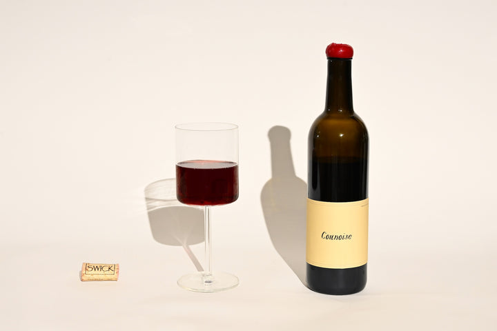 Swick Wines 2022 Counoise bottle with cream and black label, filled glass of deep red wine, and cork on beige background