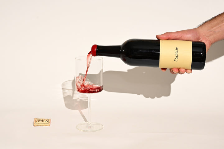 Hand pouring Swick Wines 2022 Counoise deep red wine from bottle into a glass with cork on beige background