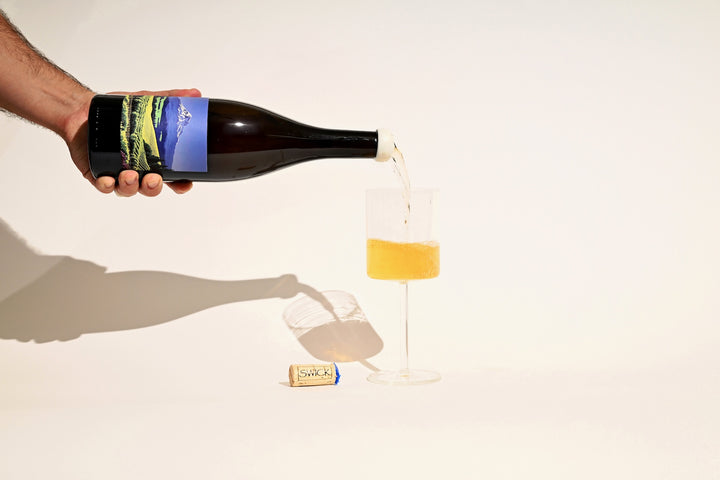 Hand pouring Swick Wines 2022 Gewurztraminer from bottle into a glass of golden amber wine with cork on beige background