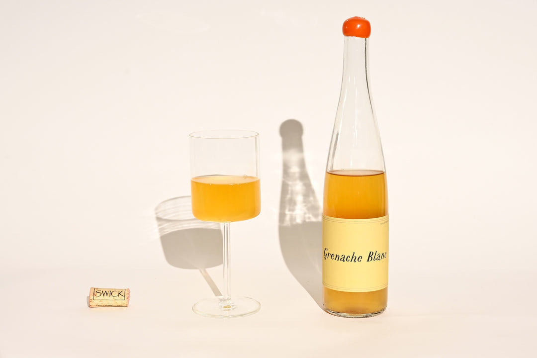 Swick Wines 2022 Grenache Blanc clear bottle with cream label, filled glass of golden amber wine, and cork on beige background
