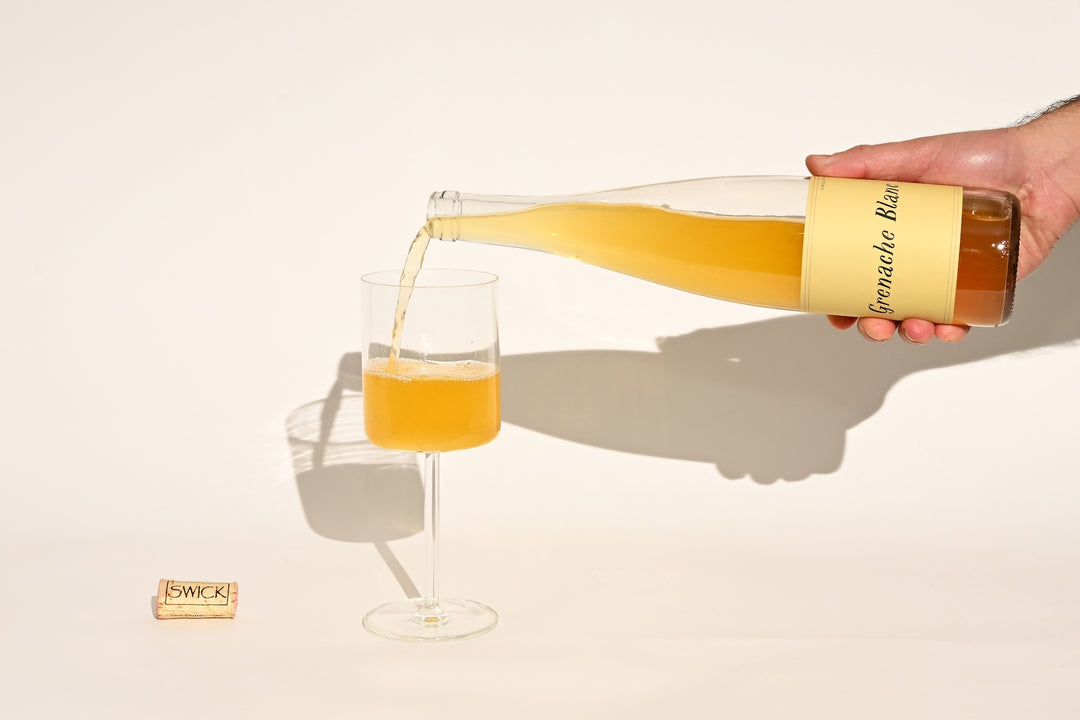 Hand pouring Swick Wines 2022 Grenache Blanc golden amber wine from clear bottle into a glass with cork on beige background