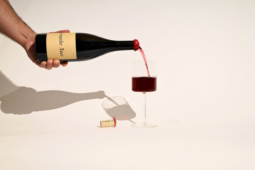 Hand pouring Swick Wines 2022 Grenache Noir red wine into a glass on beige background