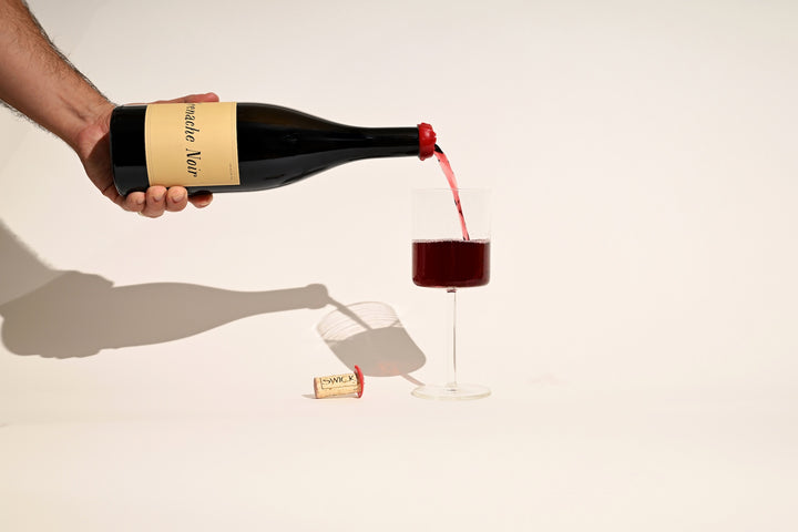 Hand pouring Swick Wines 2022 Grenache Noir red wine into a glass on beige background