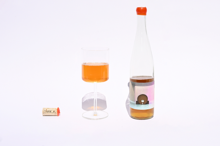 Swick Wines 2022 One Foudre clear bottle with orange cap, filled glass of amber orange skin contact wine, and small sample bottle on beige background