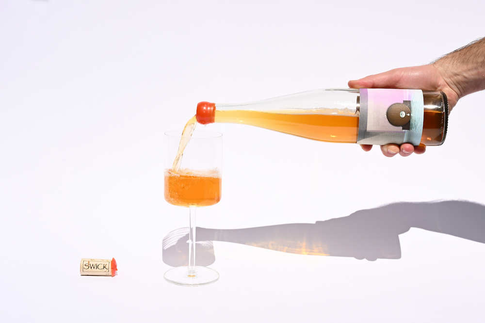 Hand pouring Swick Wines 2022 One Foudre amber orange skin contact wine from clear bottle with orange cap into a glass with cork on beige background
