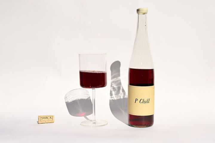 Swick Wines 2022 P-Chill clear bottle with cream and black label, filled glass of deep red wine, and cork on beige background