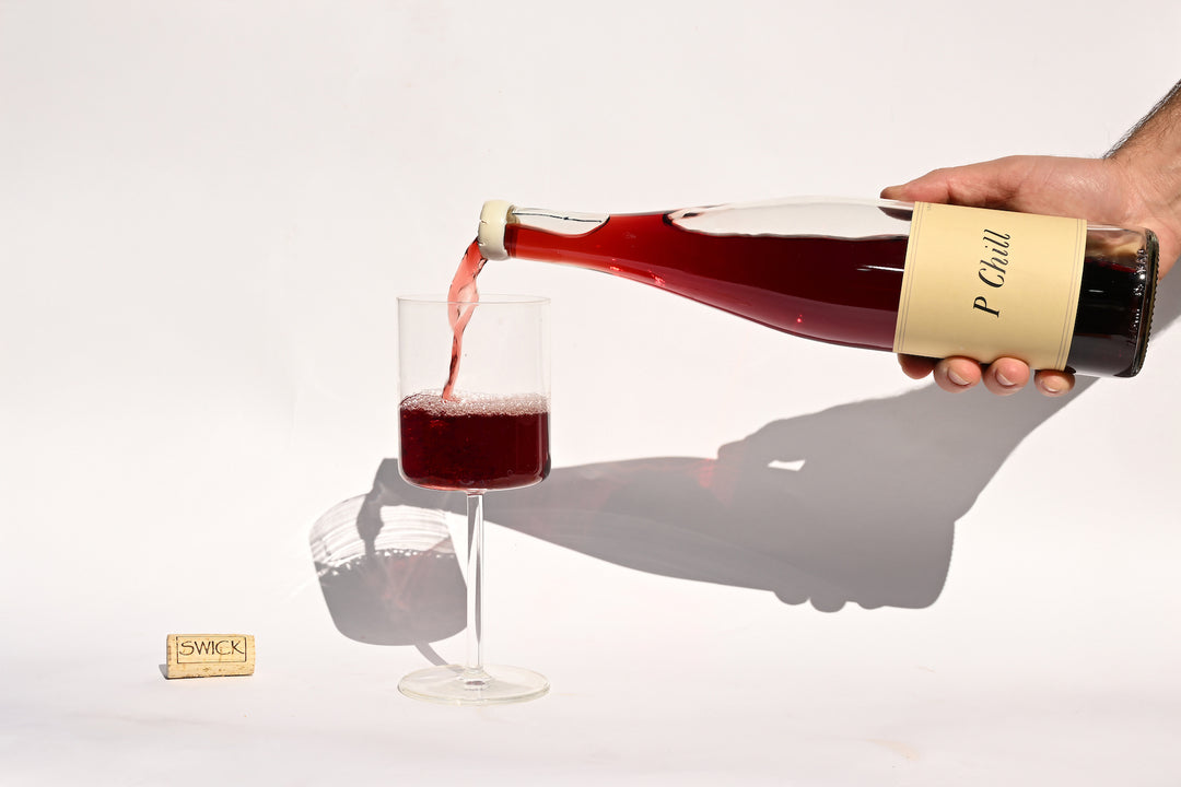 Hand pouring Swick Wines 2022 P-Chill deep red wine from clear bottle into a glass with cork on beige background