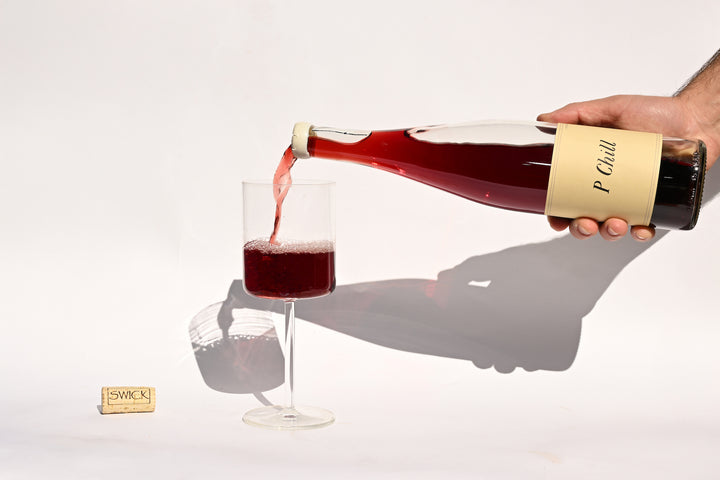Hand pouring Swick Wines 2022 P-Chill deep red wine from clear bottle into a glass with cork on beige background