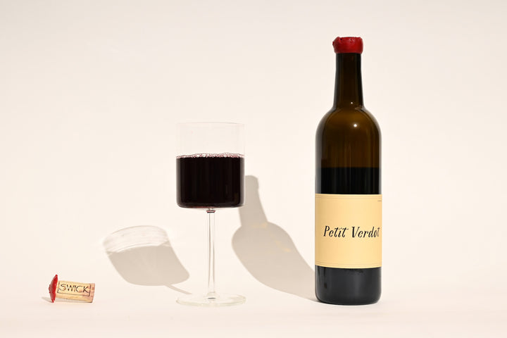 Swick Wines 2022 Petit Verdot bottle with cream and black label, filled glass of deep purple red wine, and cork on beige background