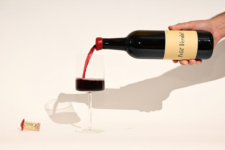 Hand pouring Swick Wines 2022 Petit Verdot deep red wine from bottle into a glass with cork and label visible on beige background