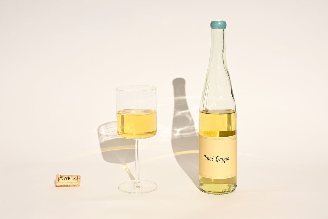 Swick Wines 2022 Pinot Grigio clear bottle with cream label and teal cap, filled glass of pale yellow white wine, and cork on beige background