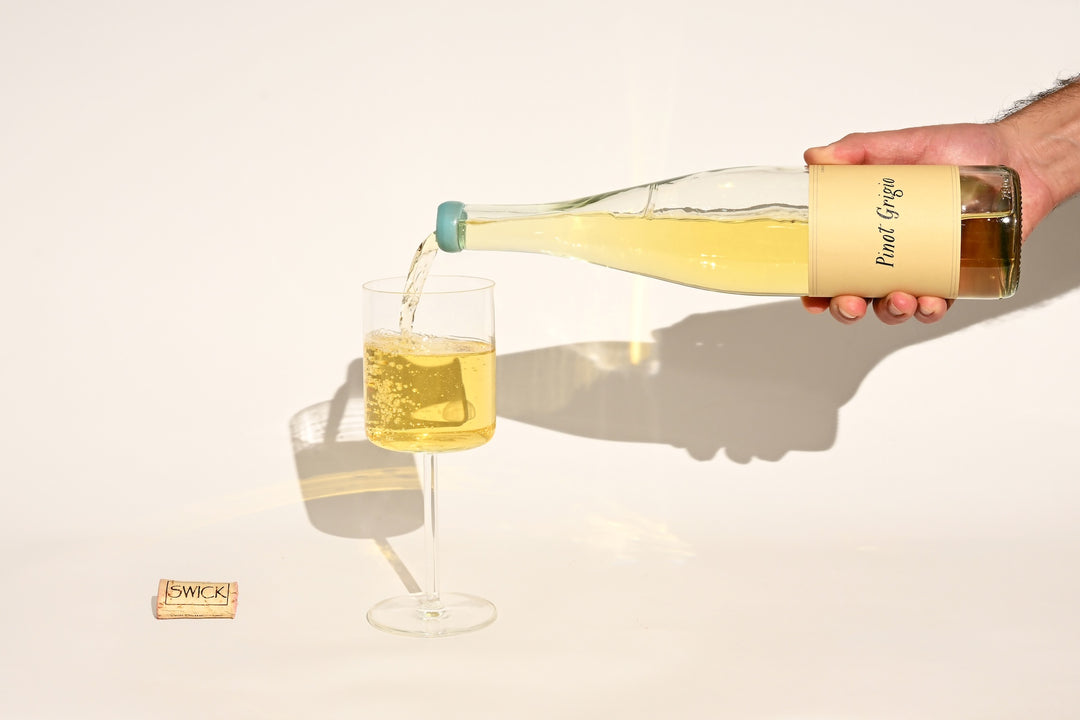 Hand pouring Swick Wines 2022 Pinot Grigio pale yellow white wine from clear bottle into a glass with cork on beige background