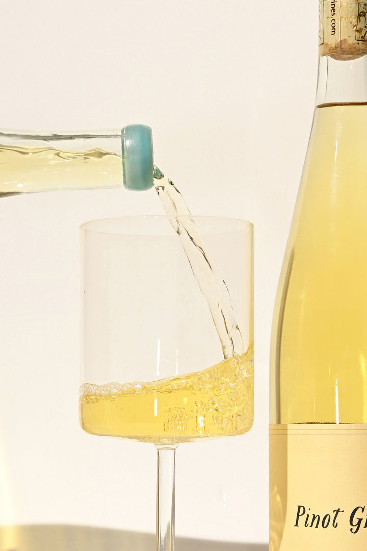 Vertical close-up of Swick Wines 2022 Pinot Grigio pale yellow wine being poured from clear bottle with teal cap into glass with label visible