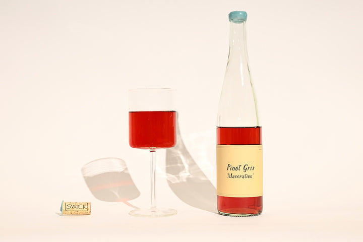Swick Wines 2022 Pinot Gris Maceration bottle with cream label, filled glass of vibrant red-pink wine, and cork on beige background