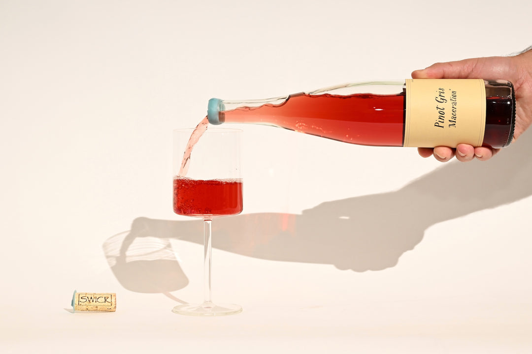Hand pouring Swick Wines 2022 Pinot Gris Maceration vibrant red-pink wine from bottle into a glass with cork on beige background