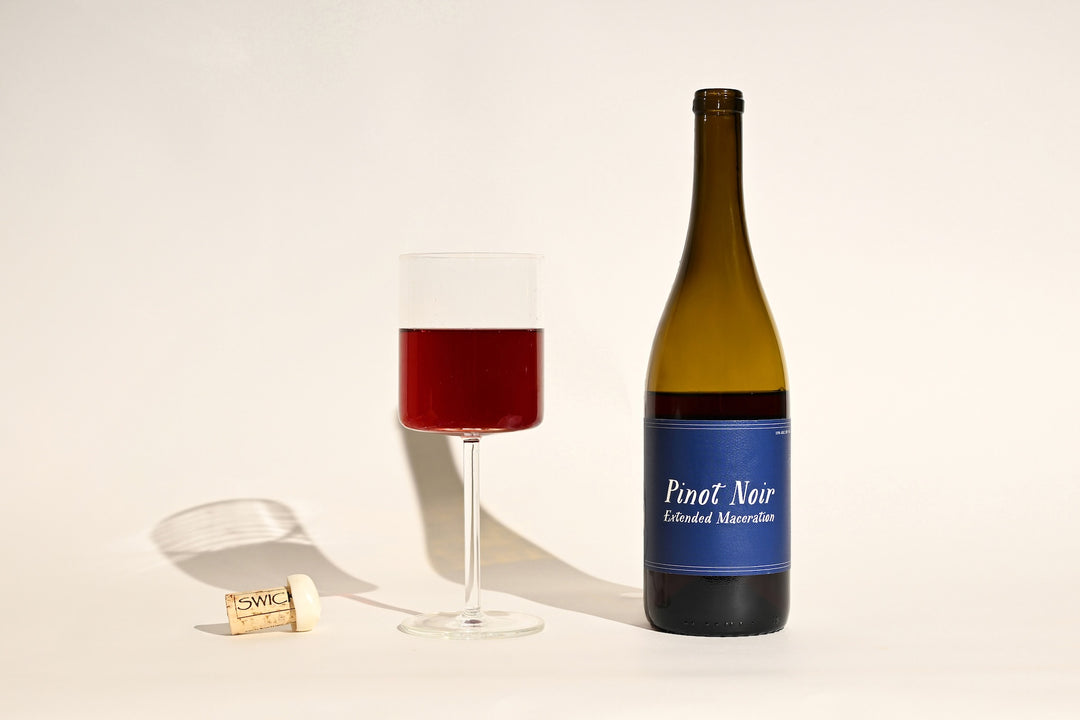 Swick Wines 2022 Pinot Noir Extended Maceration bottle with blue label, filled glass of deep red wine, and cork on beige background