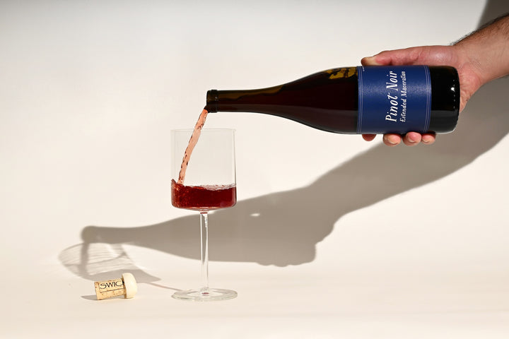 Hand pouring Swick Wines 2022 Pinot Noir Extended Maceration deep red wine from bottle into a glass with cork on beige background