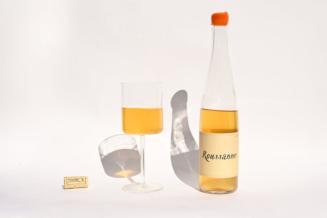 Swick Wines 2022 Roussanne clear bottle with cream label and orange cap, filled glass of golden yellow wine, and cork on beige background