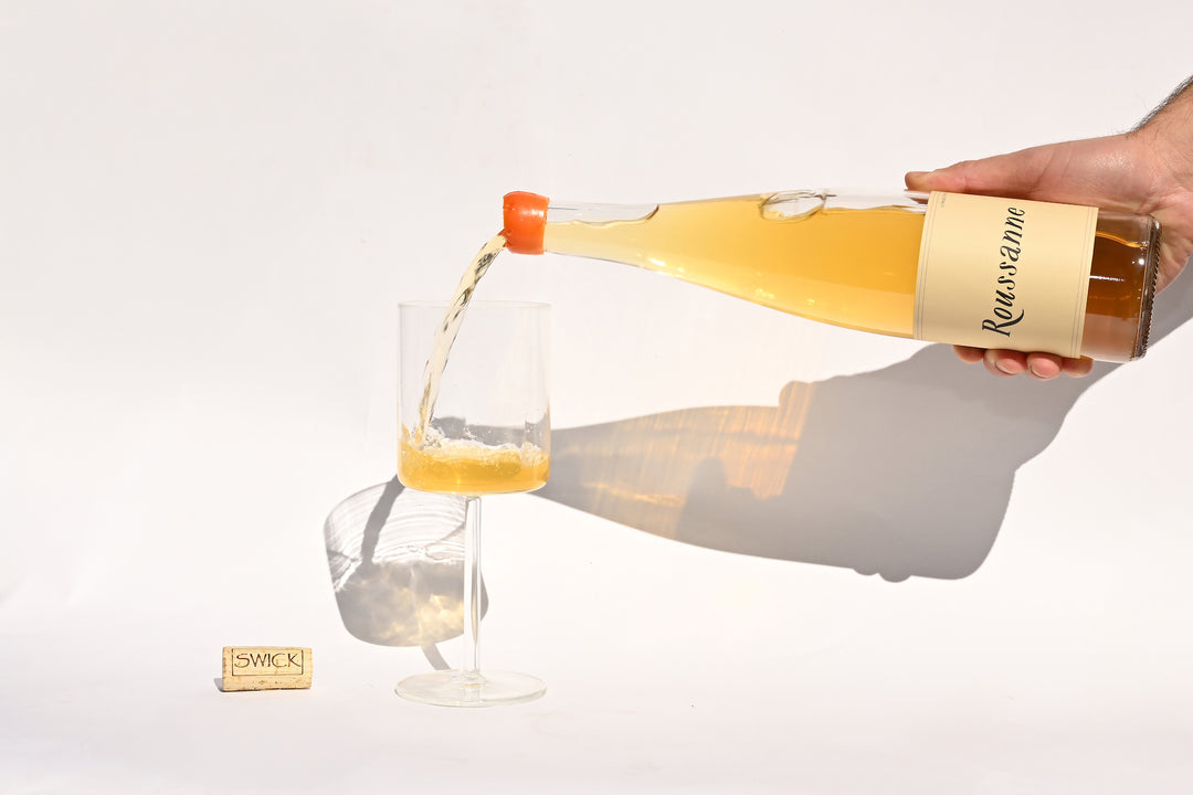 Hand pouring Swick Wines 2022 Roussanne golden yellow wine from clear bottle with orange cap into a glass with cork on beige background