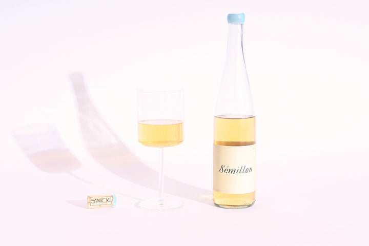 Swick Wines 2022 Sémillon clear bottle with cream label and blue cap, filled glass of golden yellow wine, and cork on beige background