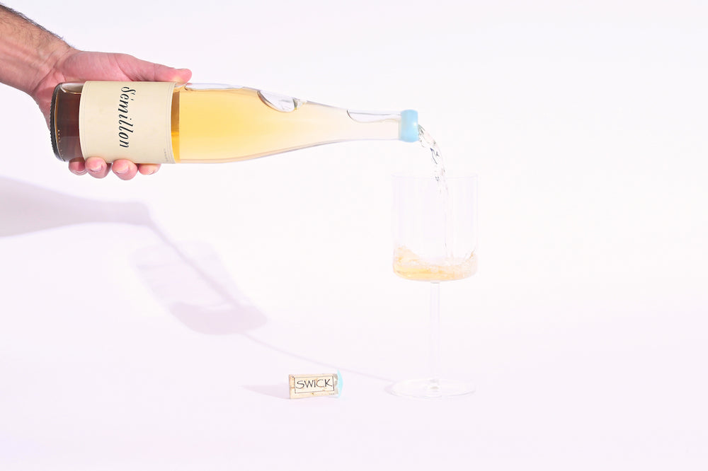 Hand pouring Swick Wines 2022 Sémillon golden yellow wine from clear bottle with blue cap into a glass with cork on beige background