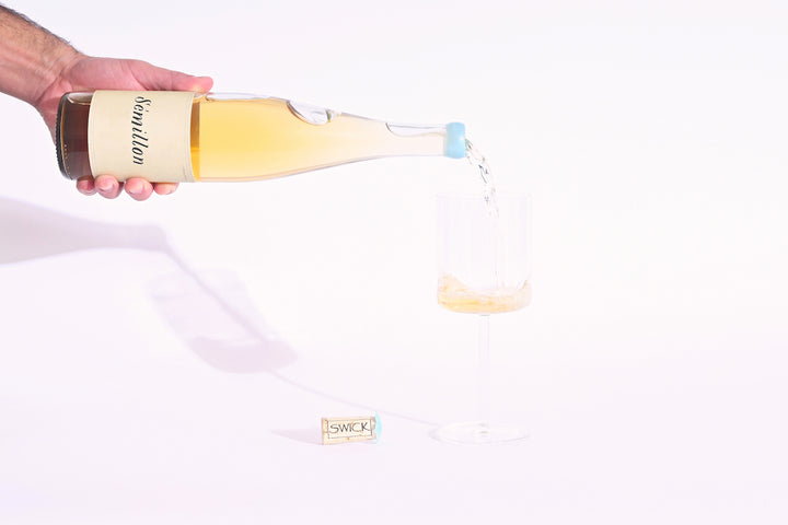 Hand pouring Swick Wines 2022 Sémillon golden yellow wine from clear bottle with blue cap into a glass with cork on beige background