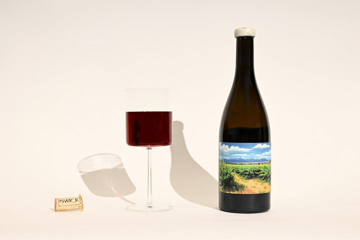Swick Wines 2022 Grenache Noir Sonrisa bottle with colorful vineyard landscape label, filled glass of deep red wine, and cork on beige background