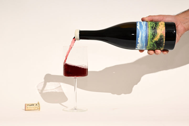 Hand pouring Swick Wines 2022 Grenache Noir Sonrisa deep red wine from bottle into a glass with cork on beige background