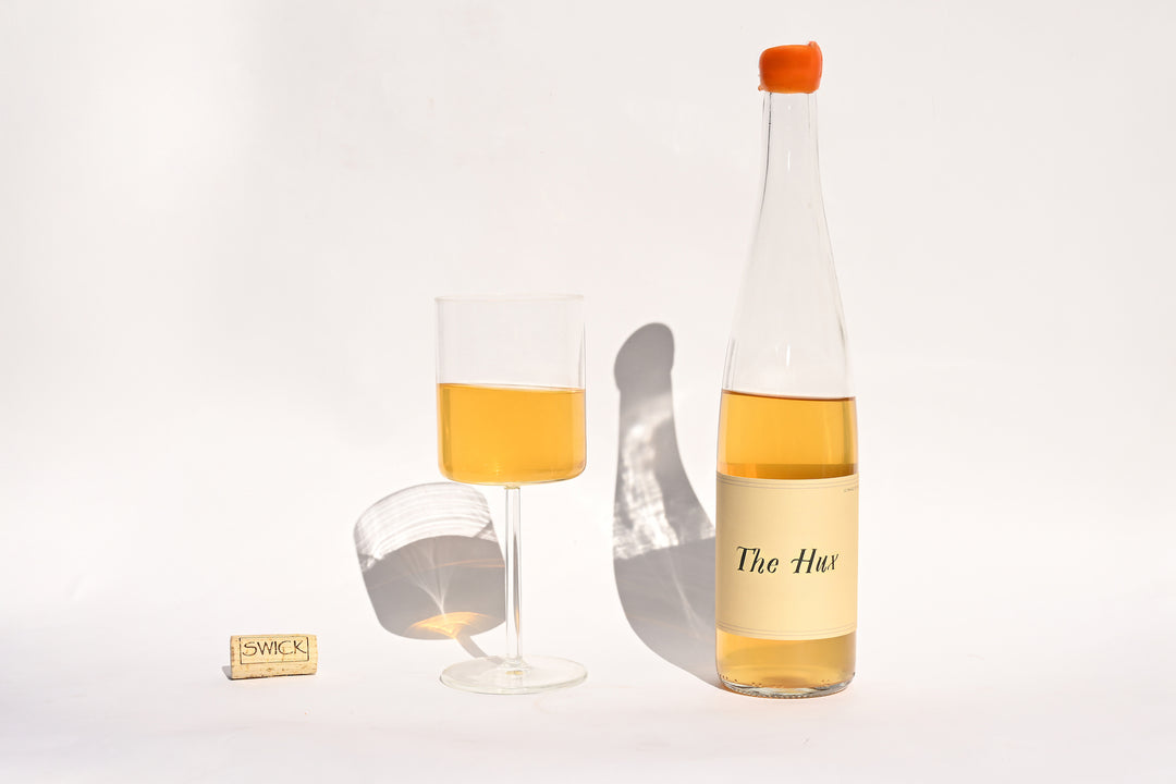 Swick Wines 2022 The Hux clear bottle with cream label and orange cap, filled glass of golden yellow wine, and cork on beige background