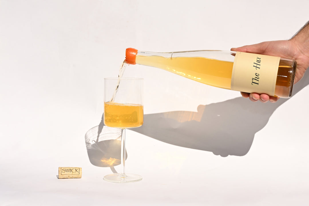 Hand pouring Swick Wines 2022 The Hux golden yellow wine from clear bottle into a glass with cork on beige background