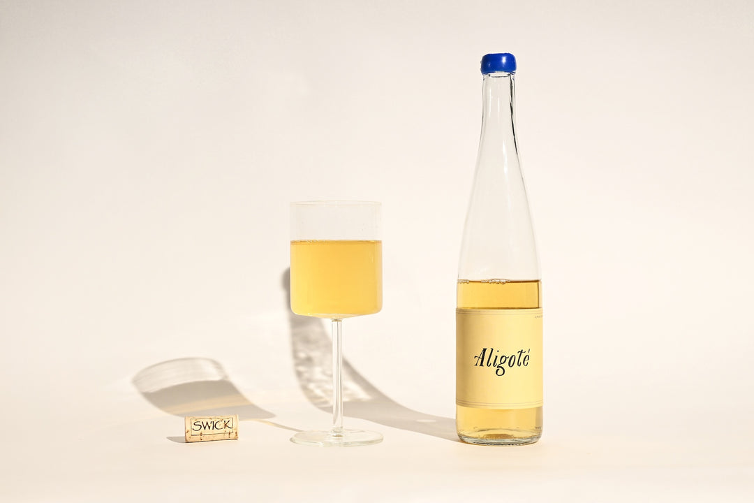 Swick Wines 2023 Aligoté bottle with cream label, filled glass of pale gold white wine, and cork on beige background