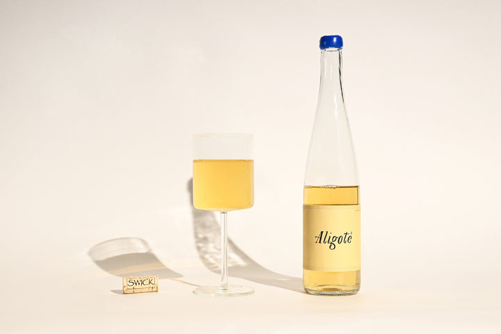 Swick Wines 2023 Aligoté bottle with cream label, filled glass of pale gold white wine, and cork on beige background