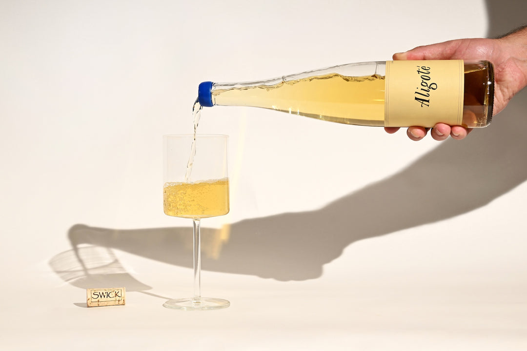 Hand pouring Swick Wines 2023 Aligoté pale gold white wine from bottle into a glass with cork on beige background