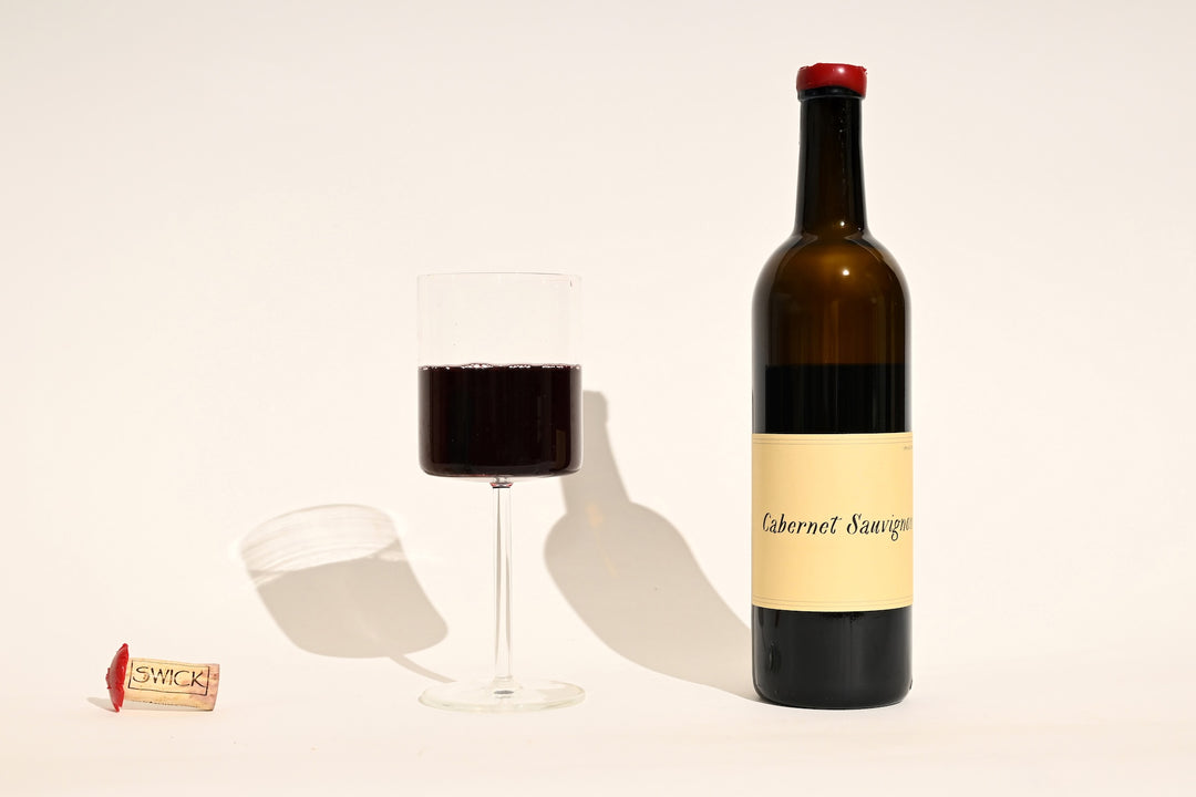Swick Wines 2023 Cabernet Sauvignon bottle with cream and black label, filled glass of deep red wine, and cork on beige background