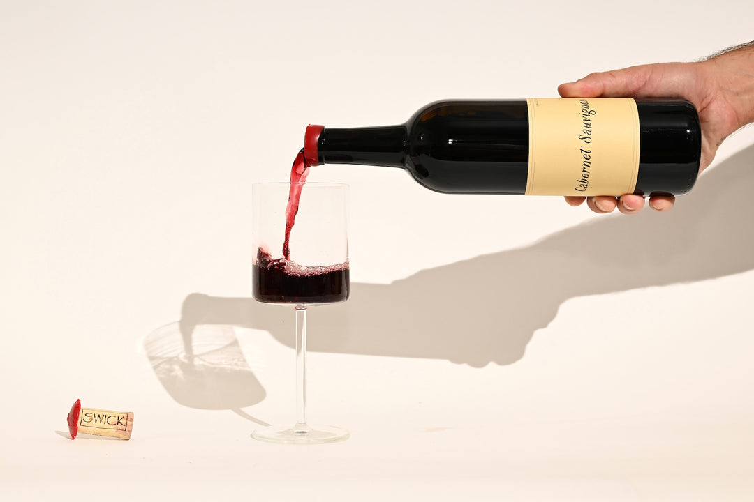 Hand pouring Swick Wines 2023 Cabernet Sauvignon deep red wine from bottle into a glass with cork on beige background