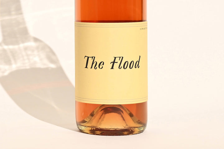 Close-up of Swick Wines 2023 The Flood bottle label with cursive text on cream background showing peach-orange rosé wine through clear glass