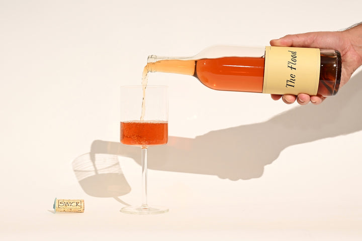Hand pouring Swick Wines 2023 The Flood peach-orange rosé wine from bottle into a glass with cork on beige background