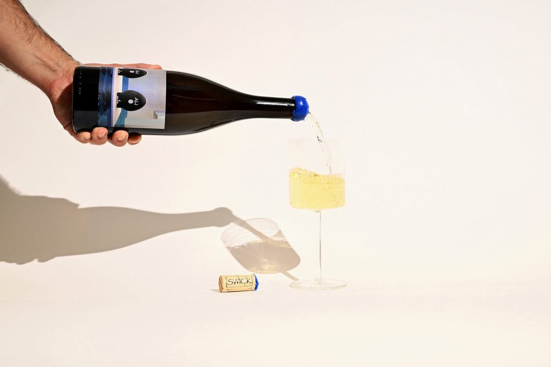 Hand pouring Swick Wines 2023 Two Eggs Aligote white wine from bottle into a glass with cork on beige background